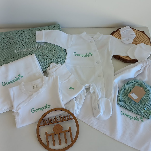 Roupa de bebé branca e verde com nome Gonçalo bordado, incluindo body, babygrow, babete, meias, gorro e manta, com placa de madeira gravada ao lado.