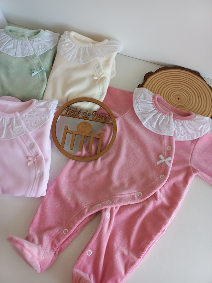 Babygrows de veludo em várias cores com golas rendadas e laços, dispostos sobre uma superfície branca com logótipo de madeira.