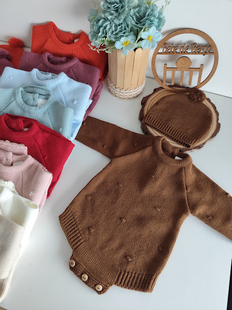 Body castanho de malha com pompons e botão, touca castanha, vários bodies coloridos, flores num vaso de madeira e placa 