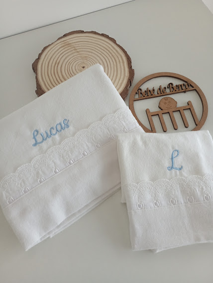 Toalhas brancas com renda e bordados azuis, base de madeira e enfeite redondo com texto Bebé de Barro
