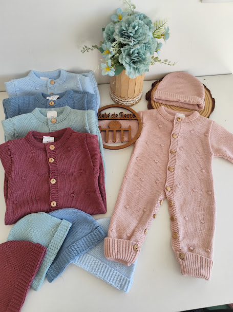 Roupa de bebé em malha colorida com textura de pequenas bolinhas e botões de madeira, incluindo macacão, casacos e gorros, e flores artificiais azuis ao fundo