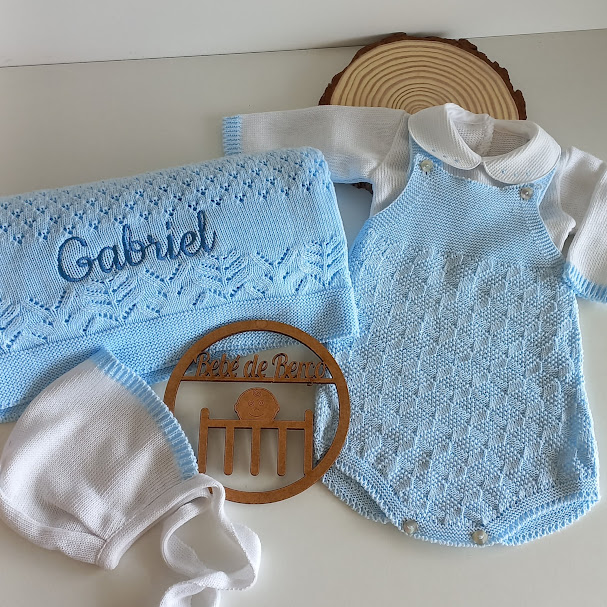Conjunto de roupa de bebé em malha azul e branca com manta bordada e touca, placa de madeira e fundo branco
