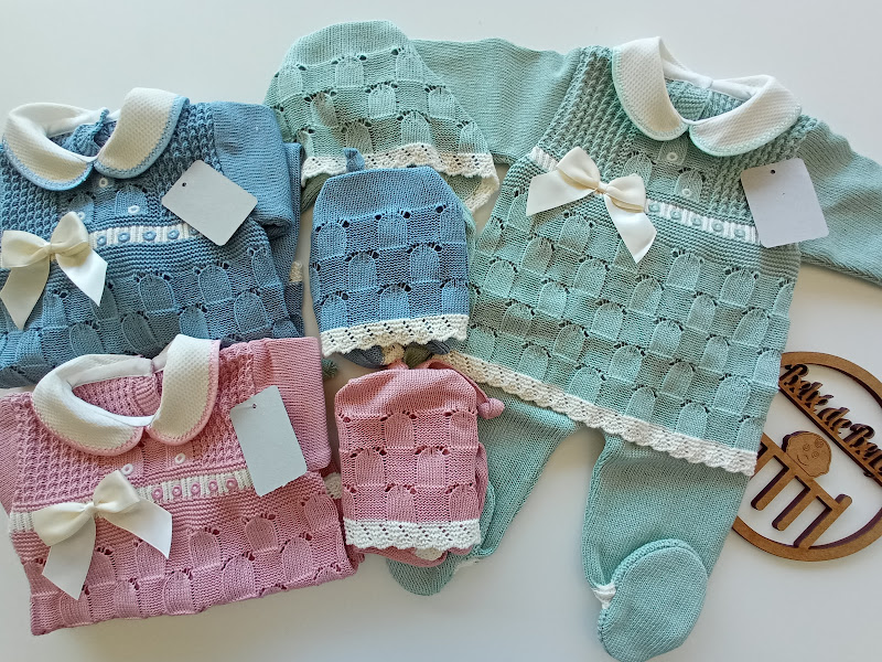 Conjuntos de roupa de bebé em tricô azul e rosa com golas brancas e laços, com etiquetas, sobre superfície clara e suporte de madeira