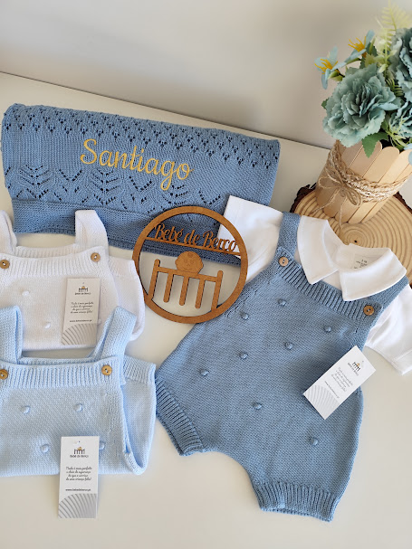 Conjunto de roupa de bebé azul e branco com manta azul bordada e ornamento de madeira em fundo com flores