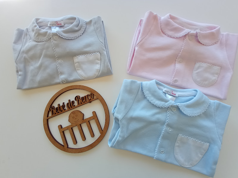 Três camisas de bebê em cores pastel com gola redonda e bolso, ao lado uma placa de madeira redonda com texto