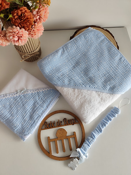 Toalha de bebé azul claro e branca, fralda branca com detalhe azul, faixa elástica azul com pendente ursinho e flores num vaso