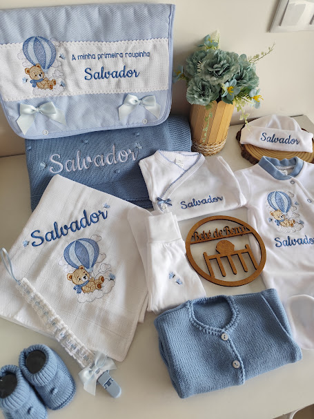 Conjunto de roupinhas de bebé azul e branco com bordados de ursinho e nome Salvador em saco armazenamento e acessórios