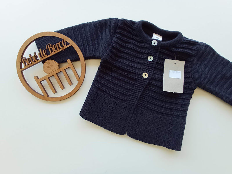 Cardigã azul-escuro de malha com botões, etiqueta pendurada, e objeto decorativo redondo de madeira com texto Arte de Barco