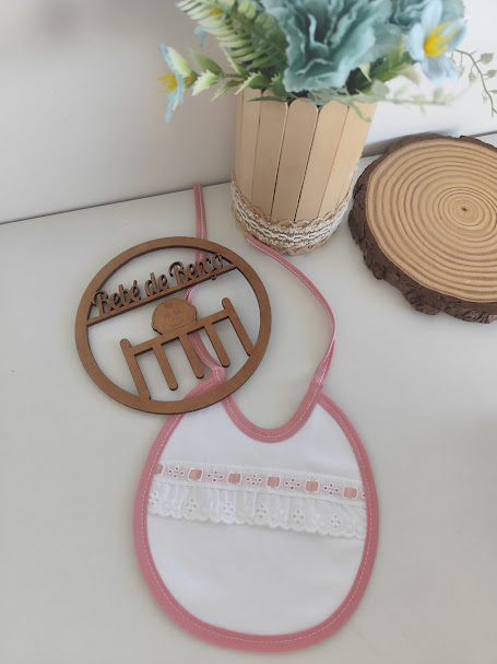 Babete de bebé branco com borda rosa e renda, enfeite de madeira redondo com texto e vaso de flores artificiais