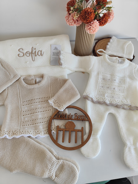 Roupa de bebé em malha branca e bege com manta personalizada, flores em vaso e decoração de madeira