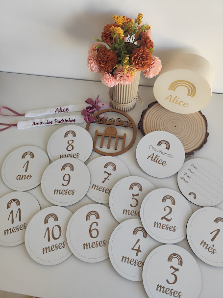 Placas brancas com meses e idades em castanho claro, flores secas em vaso creme e objetos decorativos em madeira.