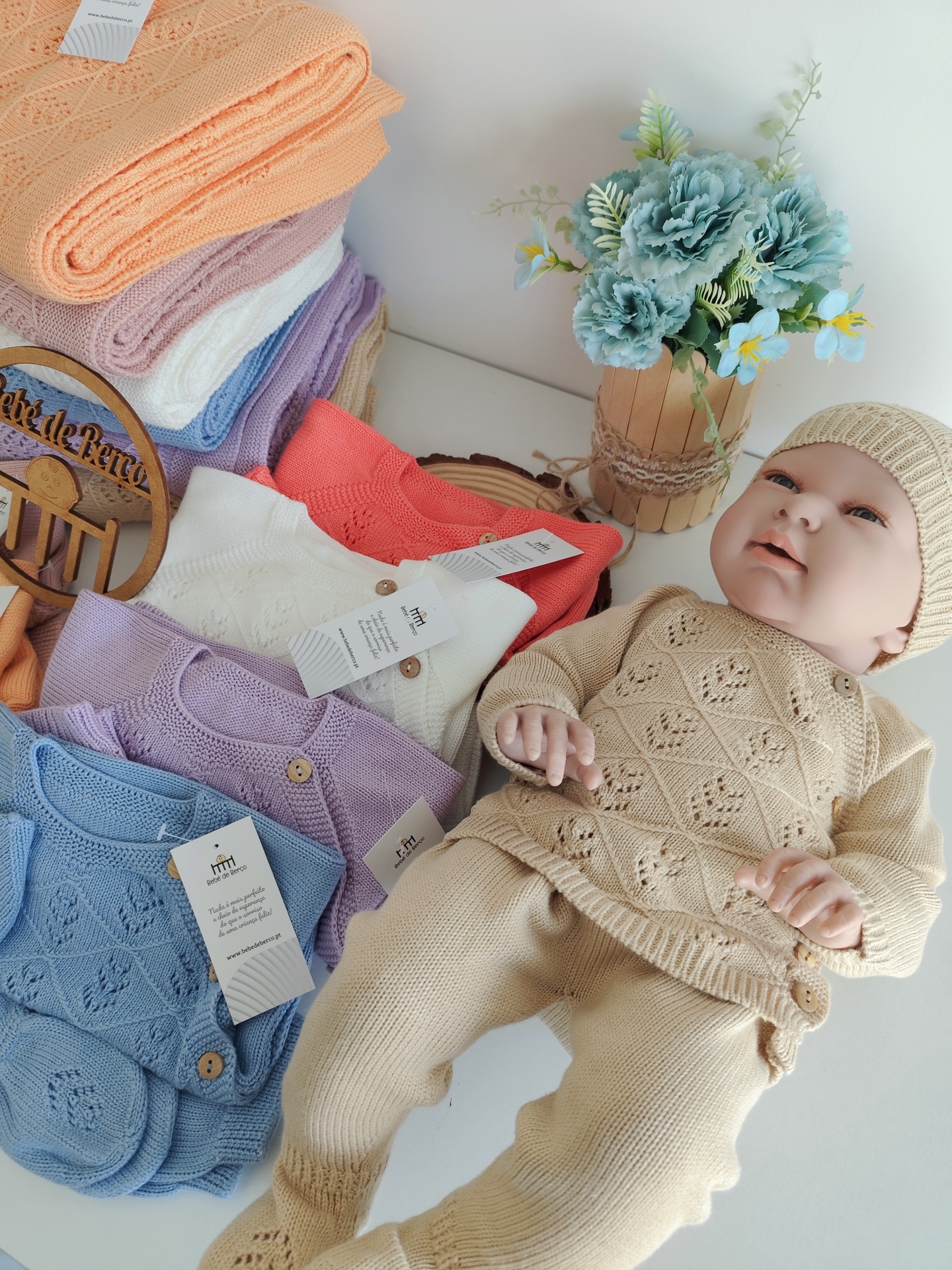 Conjuntos de roupa de bebé em malha de várias cores com padrão trabalhado, boneco vestido de creme e vaso com flores azuis