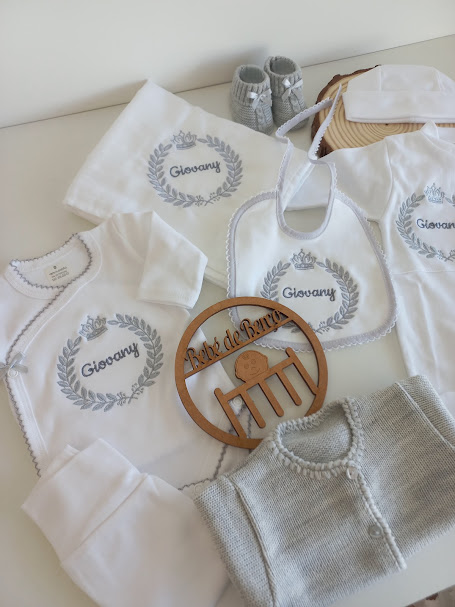 Conjunto de roupa de bebé branca com bordados cinza, acessórios de malha e marcador de madeira