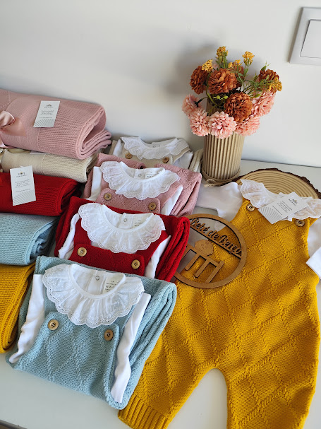 roupa de bebé em malha colorida com body branco e gola rendada, mantas e flores em vaso ao fundo