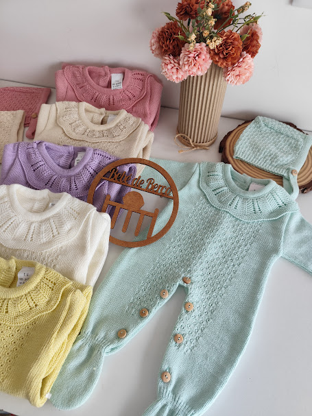 Roupas de bebé em malha colorida com botão de madeira e estampas, sobre mesa branca com flores e placa Bébé de Berço.