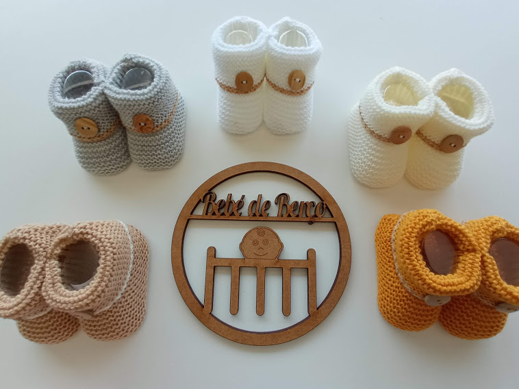 Cinco pares de sapatinhos de bebé em crochet coloridos e uma placa redonda em madeira com texto e desenho de berço