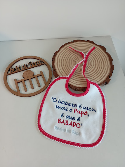 Babete branco com bordas vermelhas e texto bordado, sobre mesa branca com decorações de madeira redondas