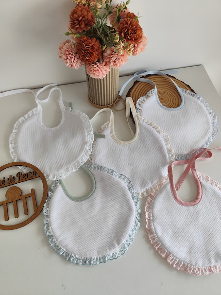 Cinco babetes de bebé com bordas de renda em várias cores e vaso com flores artificiais