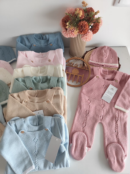 Fatos de bebé de malha em várias cores com detalhes tricotados, etiqueta e luva rosa, dispostos numa mesa branca ao lado de vaso com flores secas