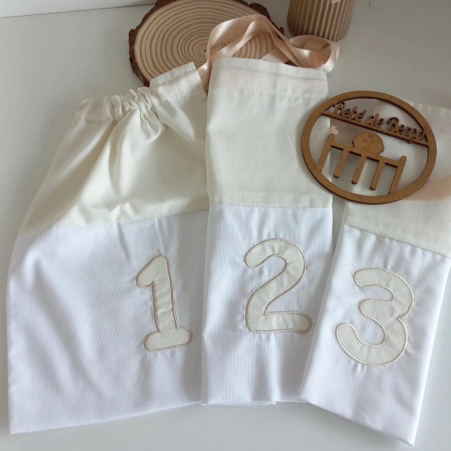 Três sacos de tecido branco e creme com números 1, 2 e 3 e uma placa de madeira redonda com texto