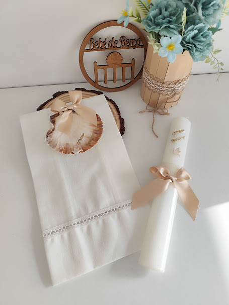 Toalha branca com renda, concha decorativa com laço rosa, vela branca com laço, vaso com flores azuis e placa de madeira