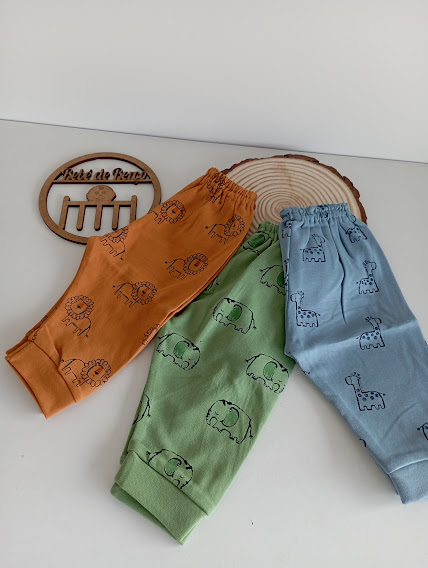 Três calças infantis coloridas com estampas de animais sobre superfície branca e objetos em madeira