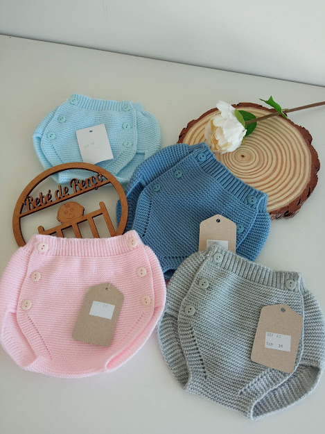 Calções de malha para bebés em azul claro, azul médio, rosa e cinzento com botões e etiquetas sobre superfície branca com flor e madeira redonda.