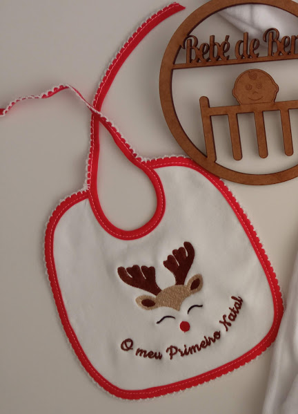 Babete branco e vermelho com rena bordada e texto, ao lado uma placa de madeira decorativa para bebé