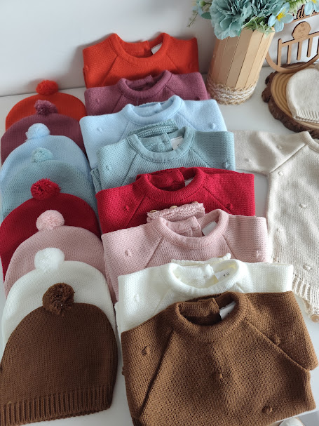 Camisolas e gorros de malha infantis coloridos organizados numa superfície branca com flores ao fundo
