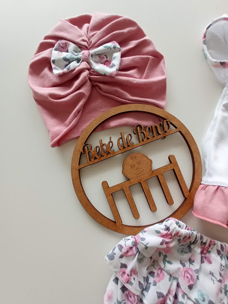 Gorro rosa com laço floral, roupa infantil branca e rosa, placa madeira 'Bebé de Banho'