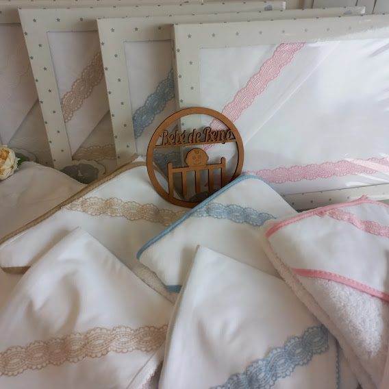 Lençóis e toalhas brancas para bebé com bordados rendados azuis, bege e rosa, embalagens transparentes e placa de madeira com texto