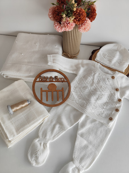 Conjunto de bebé branco em malha com casaco, calças com pés e gorro, mantas brancas, linha bege, flores e placa de madeira