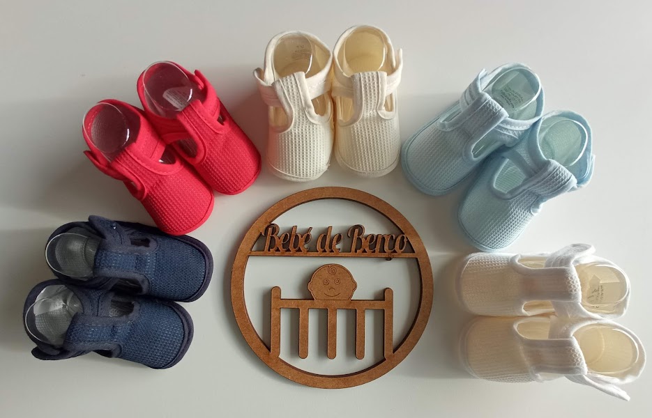 Cinco pares de sapatinhos de bebé de várias cores e um ornamento de madeira com letras e um símbolo no centro.