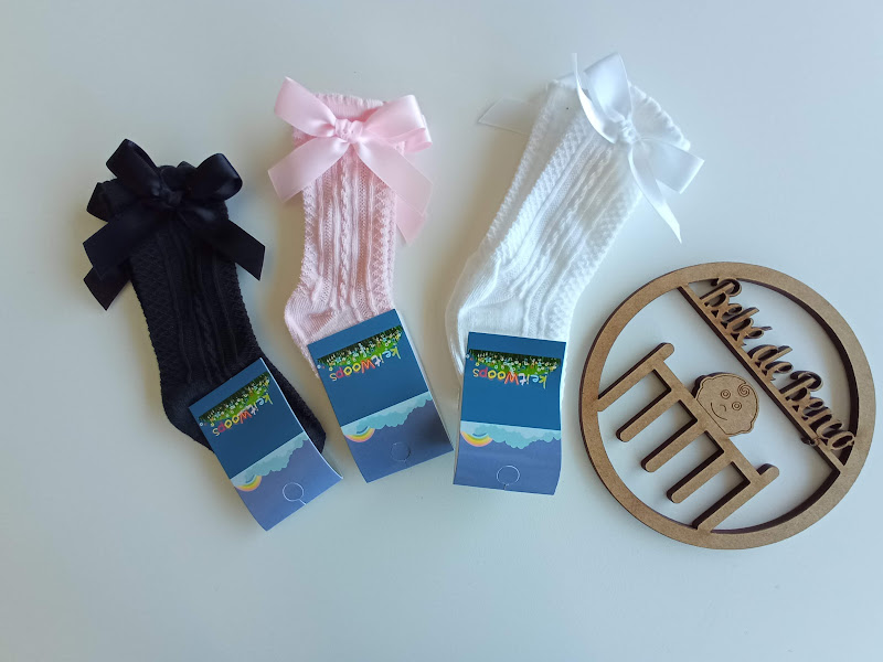 Três pares de meias para bebés com laço e etiqueta azul, ao lado placa redonda de madeira com frase e desenho