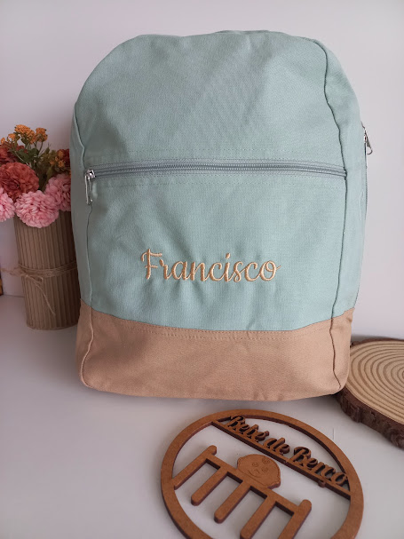 Mochila verde-água e bege com nome Francisco bordado e placa de madeira ao lado