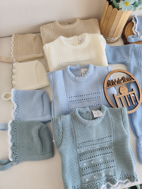 Conjunto de roupa de bebé em malha com camisolas e gorros em tons claros, detalhes bordados e enfeite de madeira