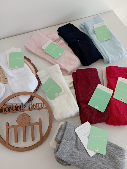 Meias coloridas para bebé com etiquetas verdes da marca 'Babybasics' sobre mesa branca, com decoração em madeira 'Bebé de Berço'
