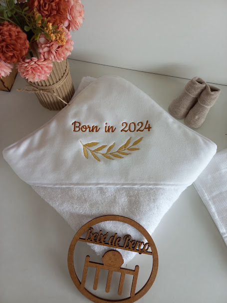 Toalha de banho branca com bordado beige 'Born in 2024', sapatos de bebé bege, flores cor de rosa e laranja, placa redonda de madeira 'Boîte de Bébé'