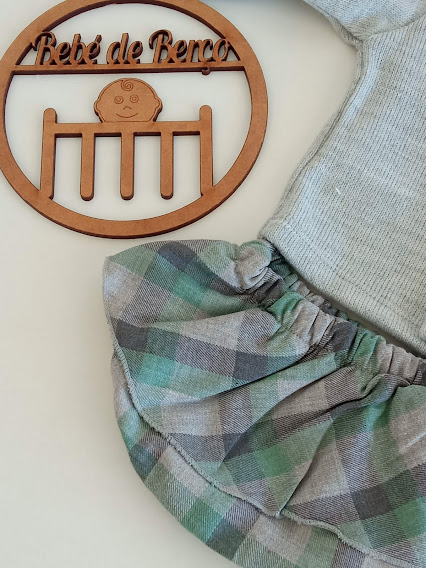 Placa decorativa de madeira com texto e desenho de bebé ao lado de roupa de bebé cinzenta com saia xadrez verde e cinza.