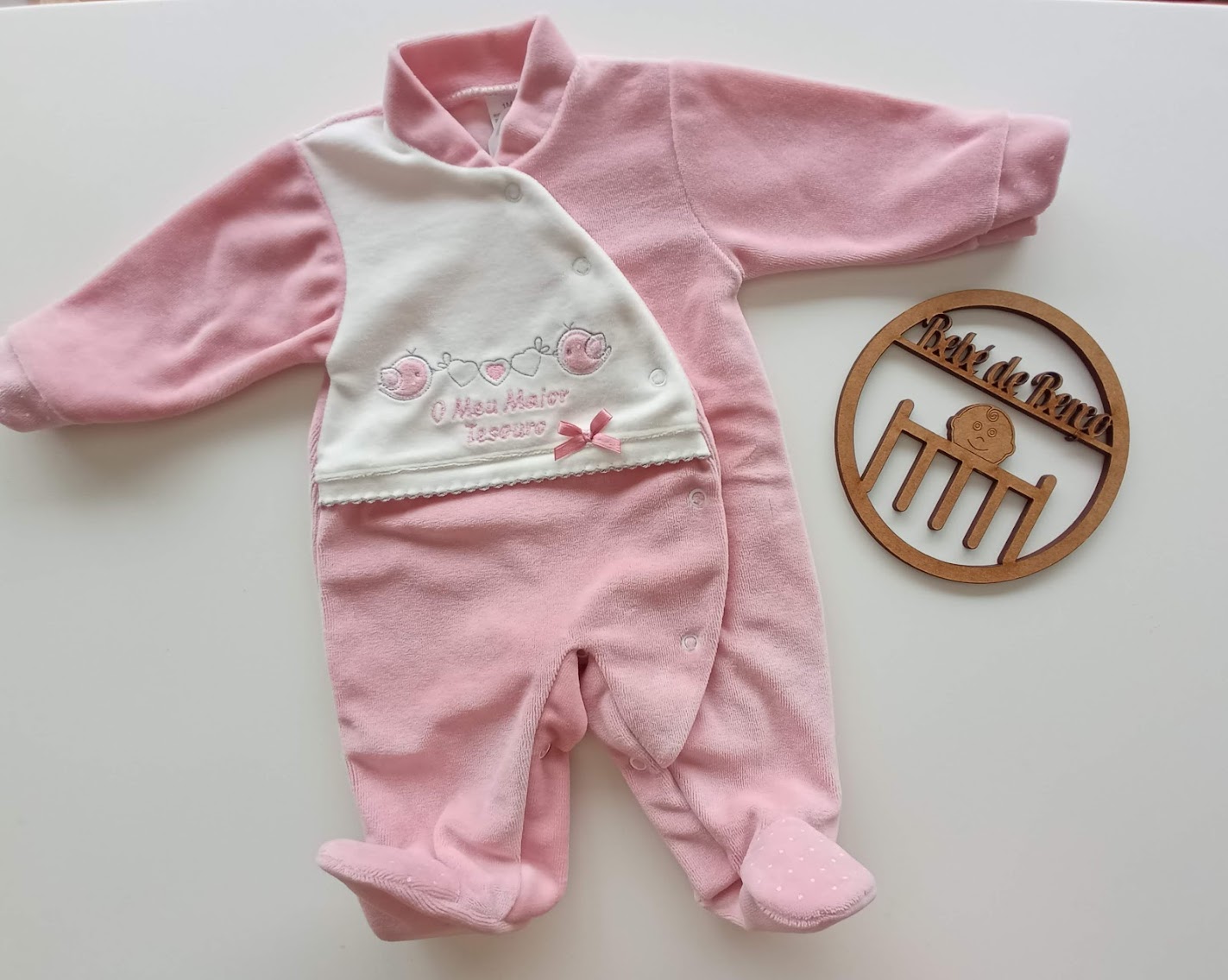 Macacão para bebé rosa e branco com bordado e fecho em diagonal colocado junto a placa de madeira redonda.