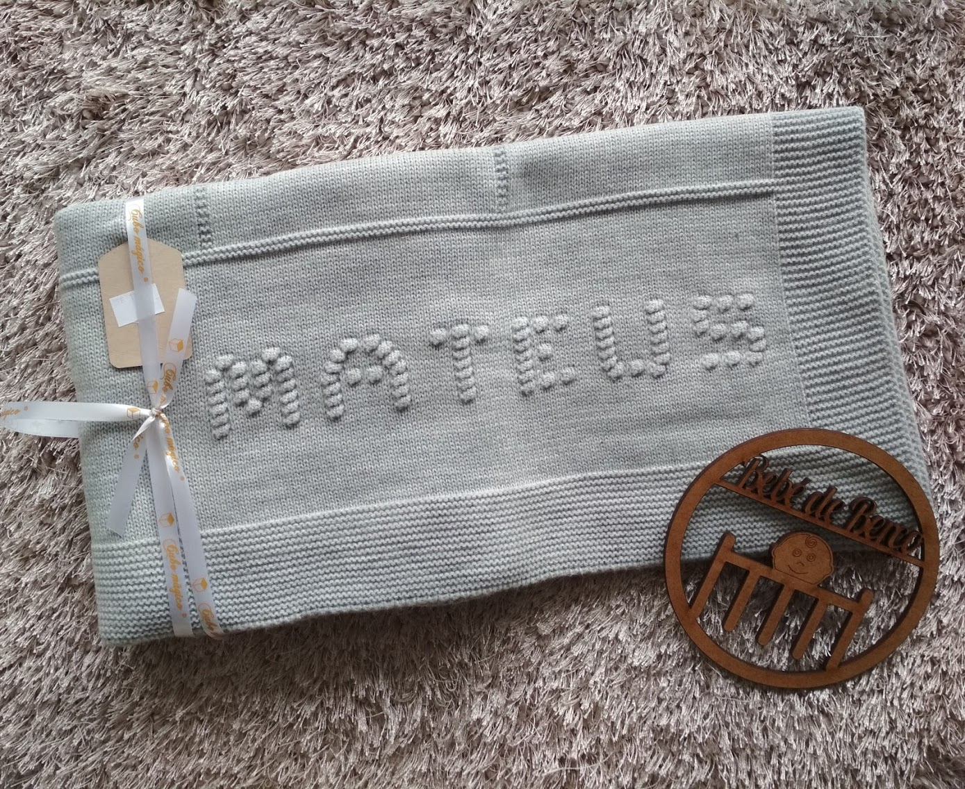 Manta tricotada cinza clara com a palavra MATEUS em relevo e enfeite de madeira ao lado