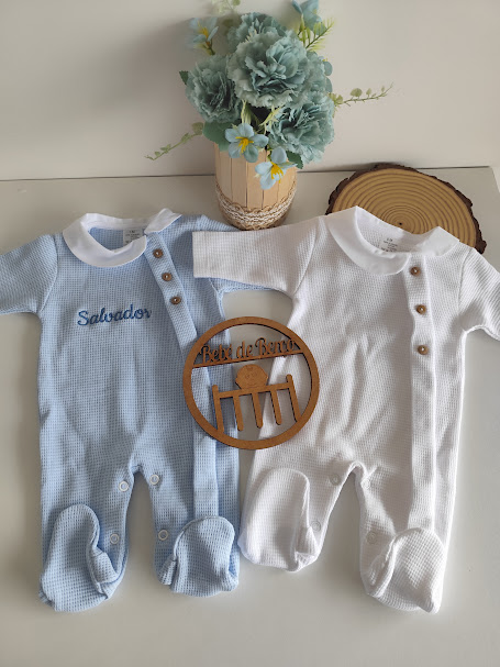 Dois macacões de bebé azul e branco com detalhes em botões junto a flores e base decorativa de madeira