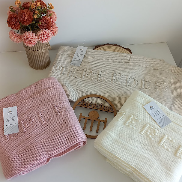 Cobertores de malha rosa, branco e bege com etiquetas, vaso de flores secas e placa de madeira numa mesa branca