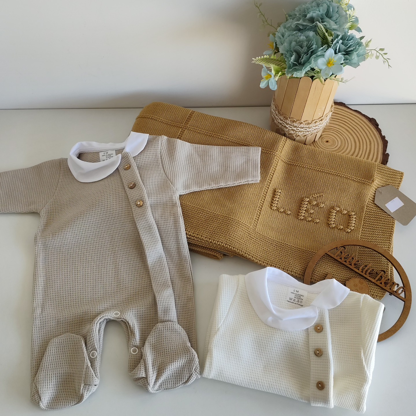 Roupa de bebé em malha bege e branca com manta castanha e vaso de flores