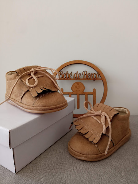 Sapatinhos castanhos em camurça com franjas e atacadores sobre caixa branca e decoração de madeira