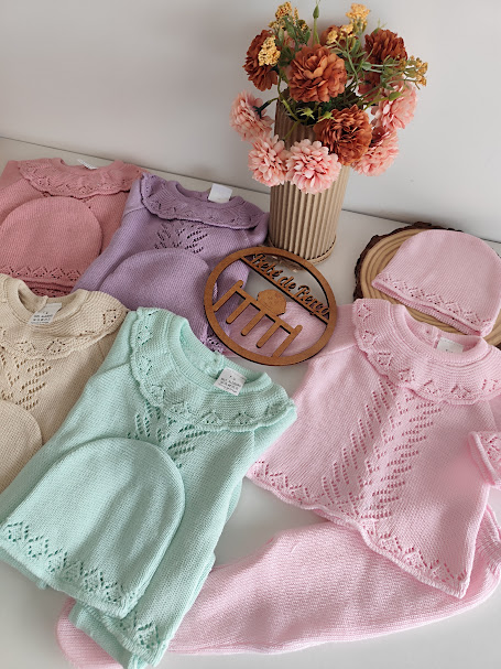 Conjunto de roupa de bebé em malha com padrões delicados em várias cores pastel, com flores em vaso ao fundo