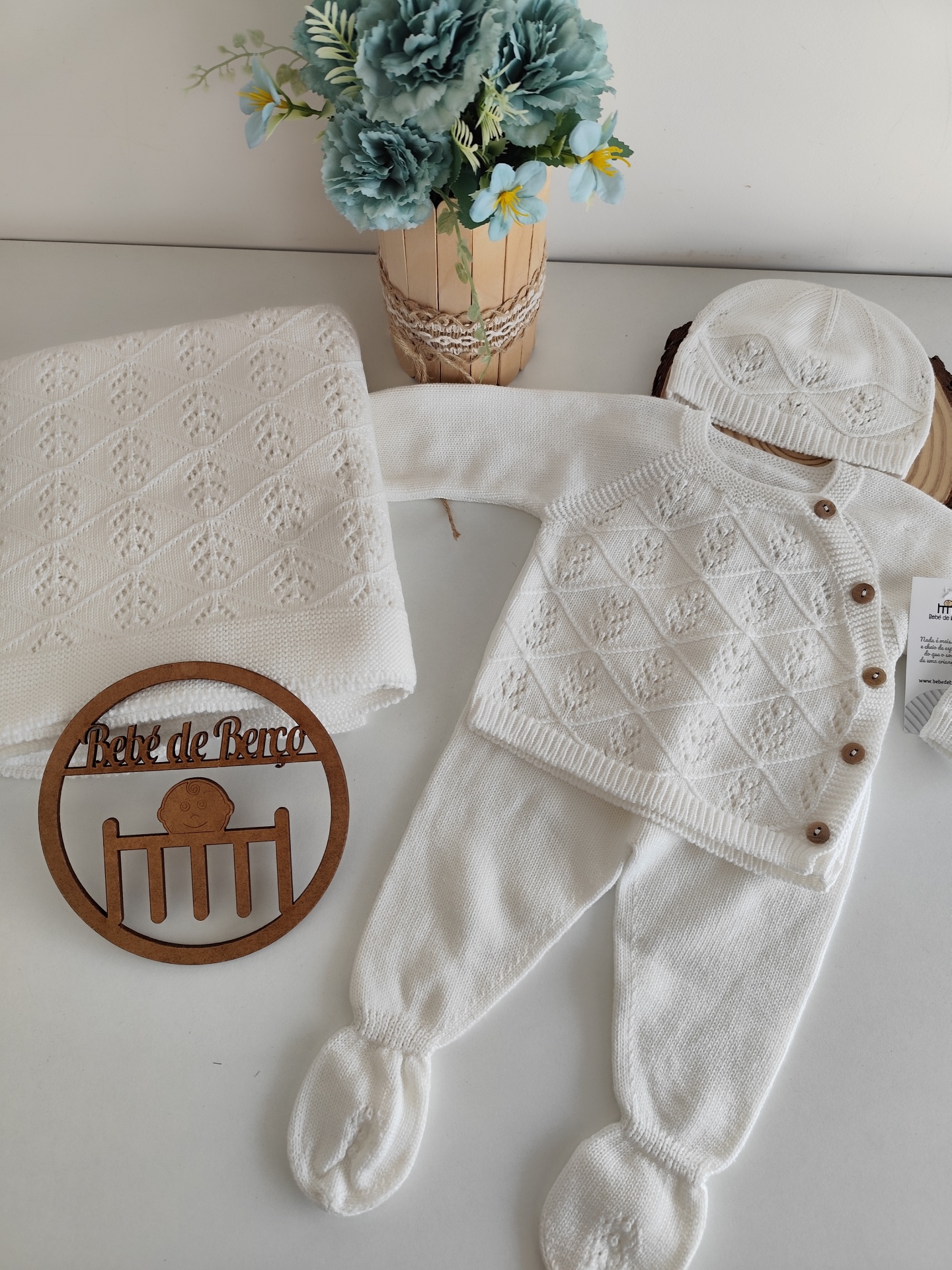 Conjunto de roupa de bebé branco em malha com padrões, manta, adorno de madeira e vaso de flores azul e amarelo