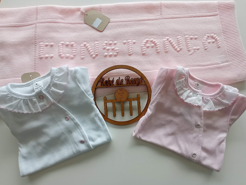 Manta rosa com letras em relevo, duas camisolas de bebé, uma branca e outra rosa, e adorno em madeira numa superfície branca