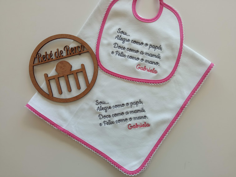 Babete e toalha brancos com bordas rosas e texto bordado ao lado de objeto redondo em madeira com texto recortado.