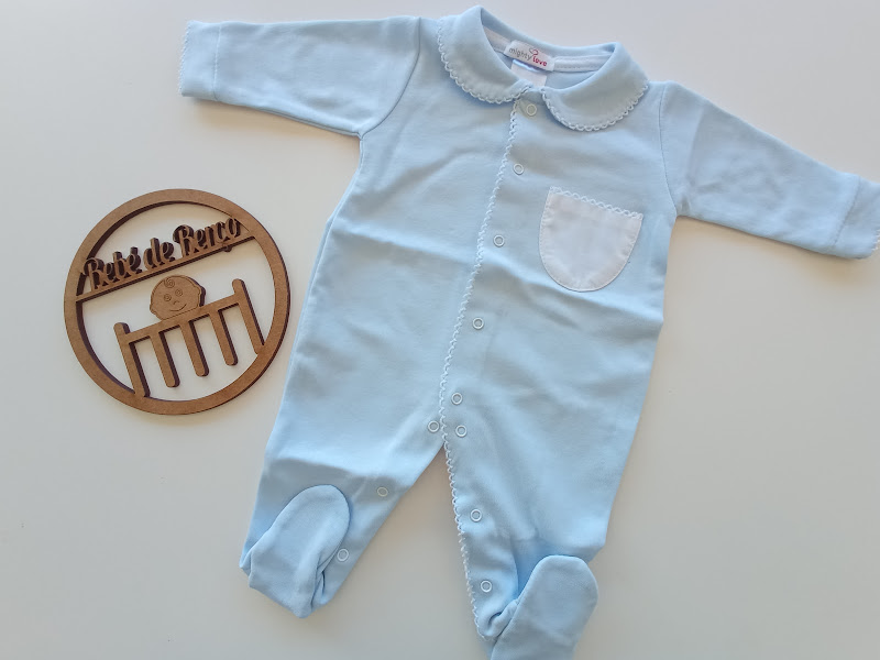Macacão de bebé azul claro com bolso branco e botões, sobre superfície branca, ao lado de placa redonda de madeira com texto